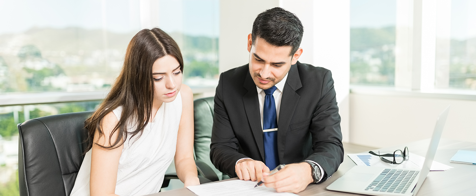 Man in suit looking over paperwork with a young woman in a brighlty lit corner office.